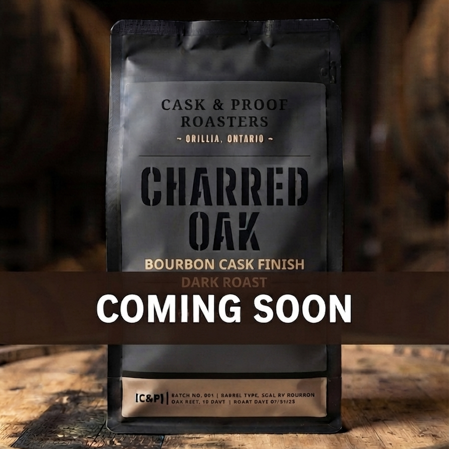 A stand-up coffee bag labeled Charred Oak Bourbon Cask Finish dark roast from Cask & Proof Roasters, photographed on a wooden barrel with a large 'COMING SOON' banner across the center.