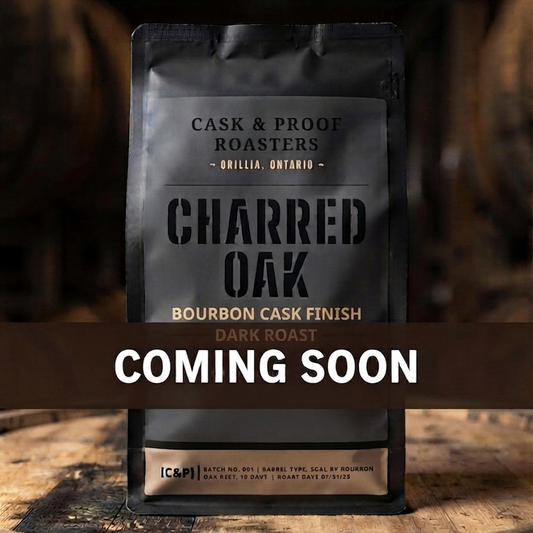 A stand-up coffee bag labeled Charred Oak Bourbon Cask Finish dark roast from Cask & Proof Roasters, photographed on a wooden barrel with a large 'COMING SOON' banner across the center.